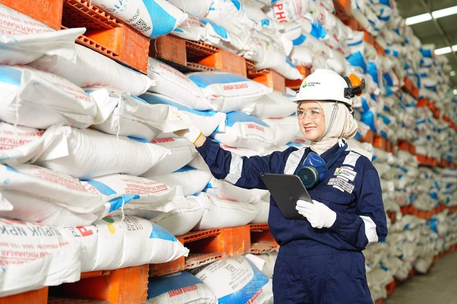 Petugas perempuan berhelm, ber-coverall, dan bersarung tangan memeriksa stok pupuk di gudang dengan tablet