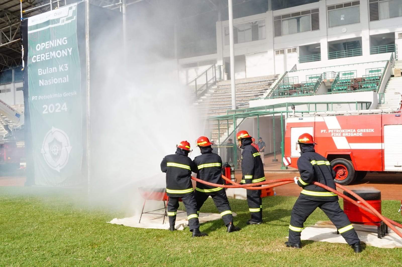Regu pemadam berseragam tahan api menyemprotkan air dalam peragaan penanganan kejadian, mobil pemadam bersiaga di belakang